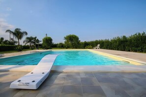 Ein Schwimmbecken mit Sonnenterrasse, umgeben von Grünpflanzen und wolkenlosem Himmel.