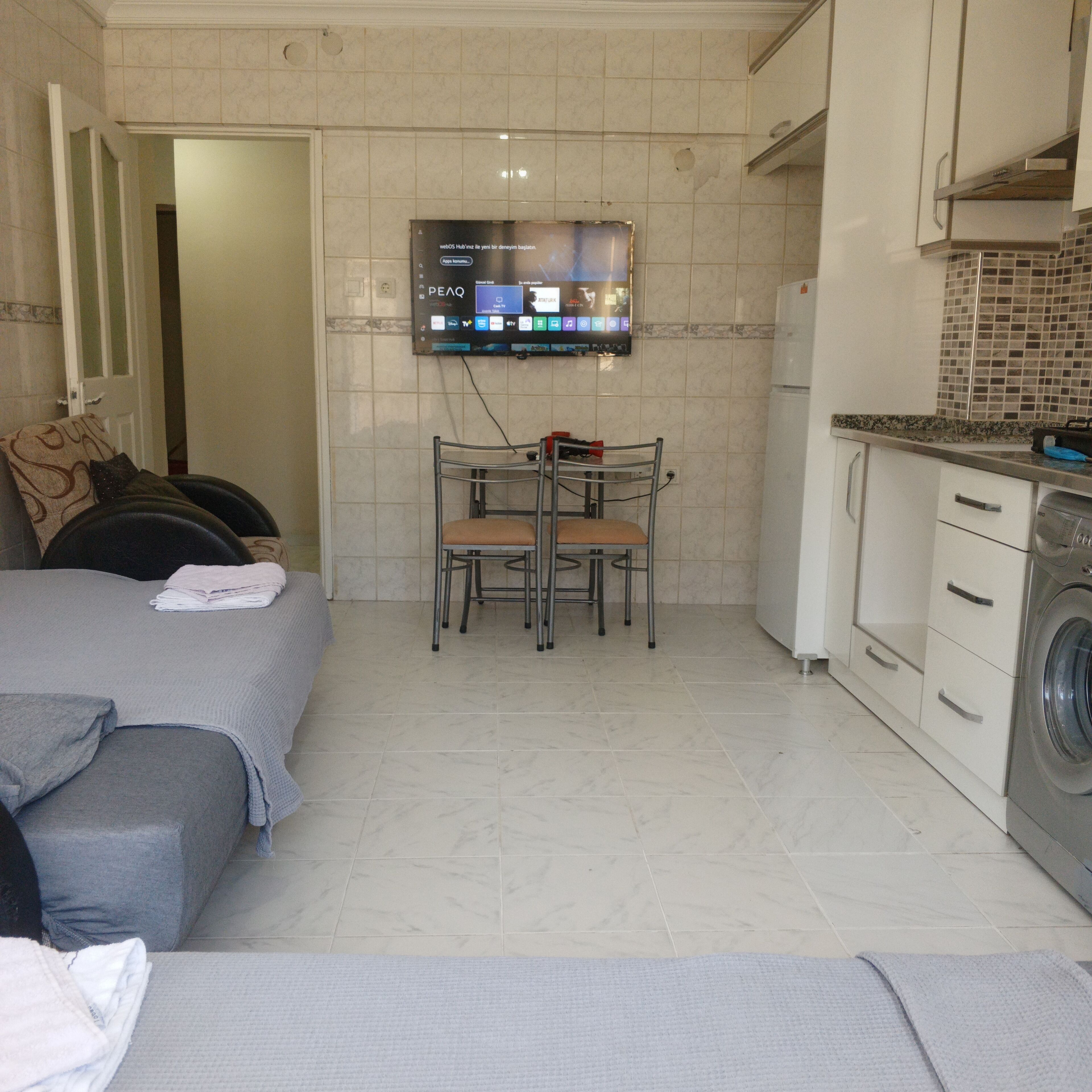A compact living space with a bed, a small table with two chairs, a TV mounted on the wall, and a washing machine.