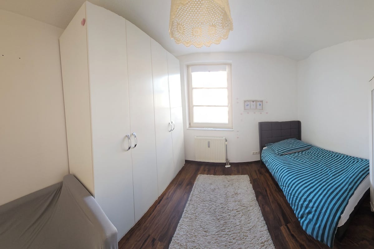 A small, well-lit room with a bed, a radiator, a window, and a wardrobe.