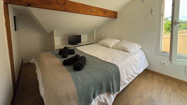 Un dormitorio con techo inclinado, una cama con ropa de cama blanca, una manta verde de punto y un televisor sobre un estante blanco.