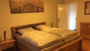 Una cama doble con estructura y cabecera de madera, una mesita de noche con lámpara, un cuadro de un muelle en la pared, y una puerta con cortina.