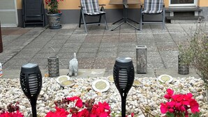 Un patio al aire libre con dos sillas, una mesa y un pequeño jardín con flores rojas e iluminación decorativa.