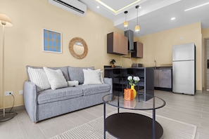 A modern living room with a grey sofa, a glass coffee table, and a kitchen area with cabinets and a refrigerator.