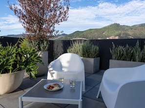 A rooftop terrace with white furniture, a dining table with a plate of food and drinks, and potted plants.