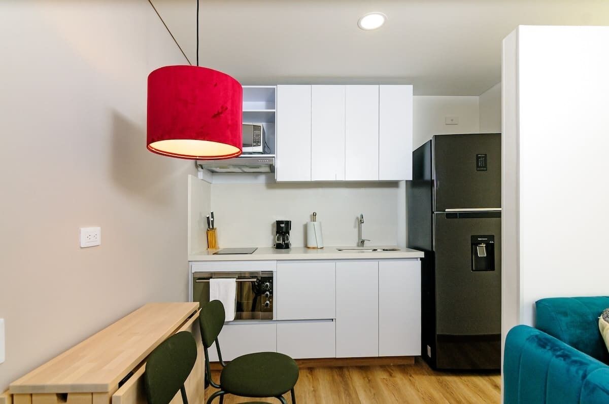 Une cuisine compacte avec des meubles blancs, un réfrigérateur en acier inoxydable, un luminaire suspendu rouge, une table en bois et un canapé de couleur turquoise.