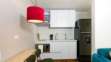 Une cuisine compacte avec des meubles blancs, un réfrigérateur en acier inoxydable, un luminaire suspendu rouge, une table en bois et un canapé de couleur turquoise.