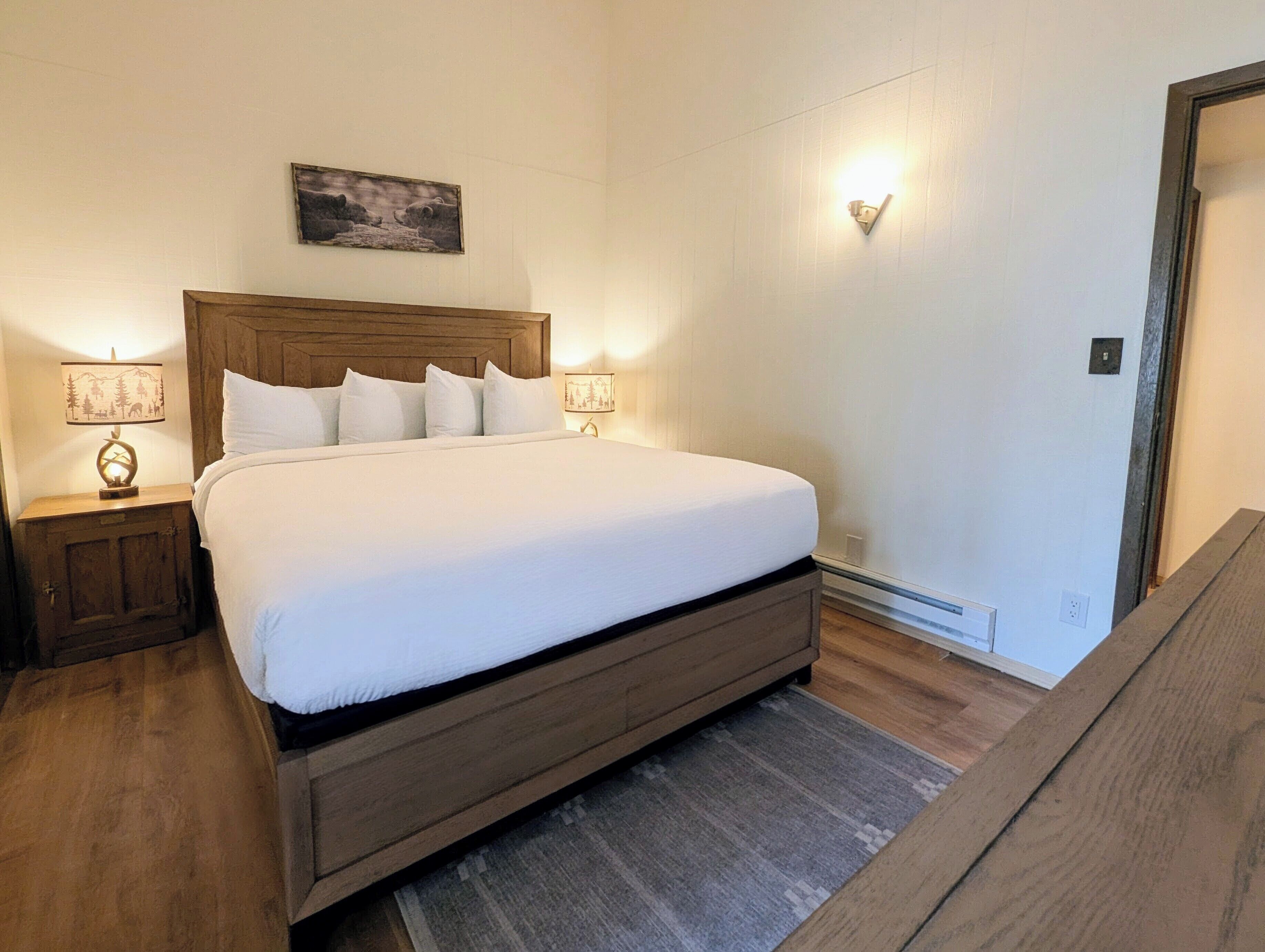 Ein ordentlich bezogenes Bett in einem Hotelzimmer, mit einem hölzernen Kopfteil, zwei Nachttischen mit Lampen und einem gerahmten Bild an der Wand.
