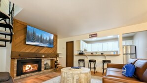 A living room with a fireplace, a round ottoman, a television, and a kitchen area with bar stools. - Peaceful Retreat.  Outdoor Pool / Indoor Hot Tub (Keystone)