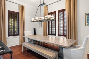 A dining area with a wooden table and benches, large windows with shutters, and a hanging light fixture.
