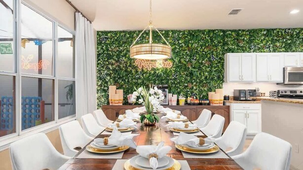 Un comedor con una larga mesa de madera dispuesta para una comida, sillas blancas y un letrero de neón en la pared.