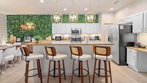 Una cocina moderna con armarios blancos, una isla central con taburetes altos, un letrero de neón y una pared verde con plantas.