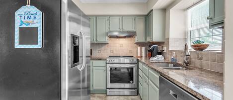 Cozinha com eletrodomésticos de aço inoxidável, armários verdes, painel de azulejos e janela com persianas.