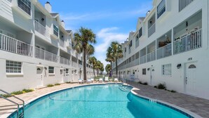A swimming pool surrounded by white buildings with balconies, palm trees, and a clear sky.