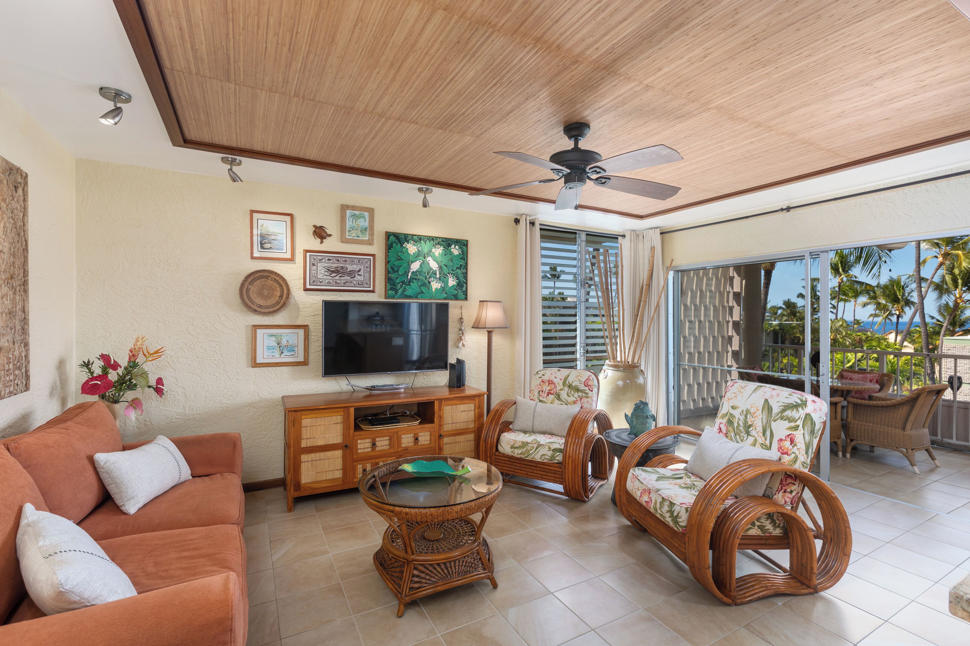A spacious living area with a television, wicker furniture, and a balcony with a view of palm trees.