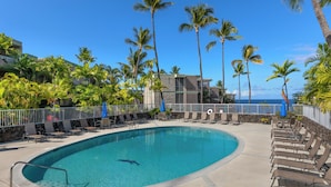 A swimming pool with lounge chairs and palm trees, surrounded by a stone wall and a building with a balcony.
