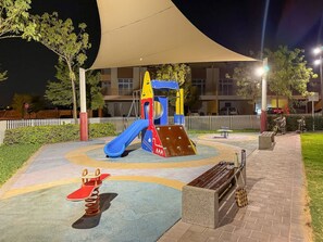 A playground with a slide and climbing structure, surrounded by a paved area and a building in the background.