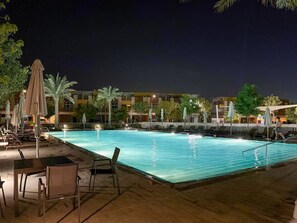 Uma área de piscina à noite, com espreguiçadeiras e guarda-sóis, cercada por um edifício com várias varandas.