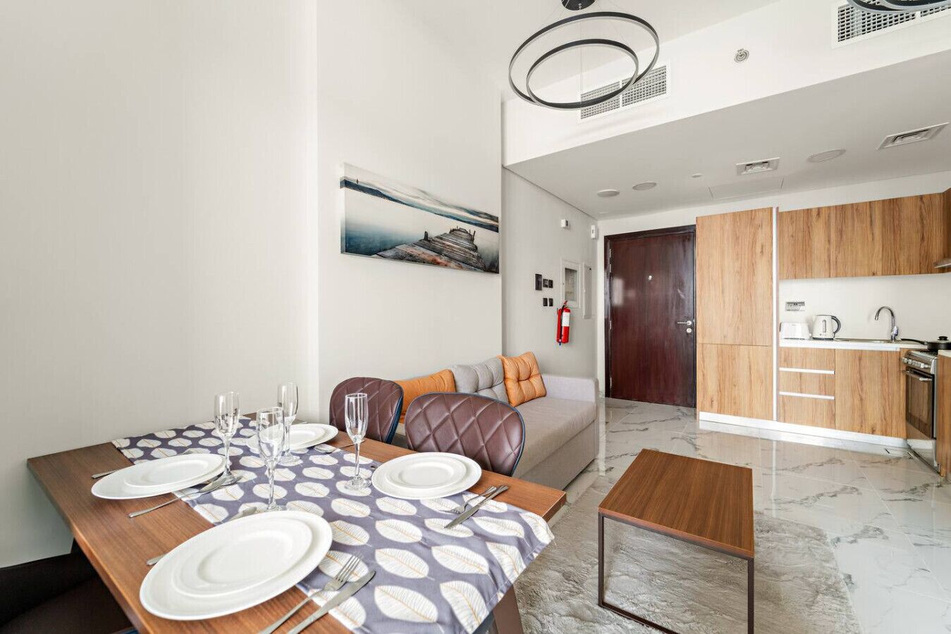 Un comedor moderno con una mesa preparada para cuatro, un sofá con cojines, una cocina con armarios de madera y un cuadro en la pared.