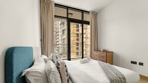 Une chambre avec un grand lit, une chaise bleue, une commode en bois et une vue sur un immeuble doté de balcons.