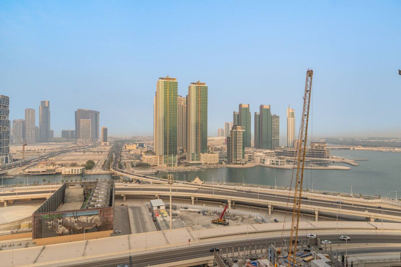 Un paysage urbain avec des immeubles modernes de grande hauteur, une rivière et une autoroute où l’on aperçoit des grues de chantier.