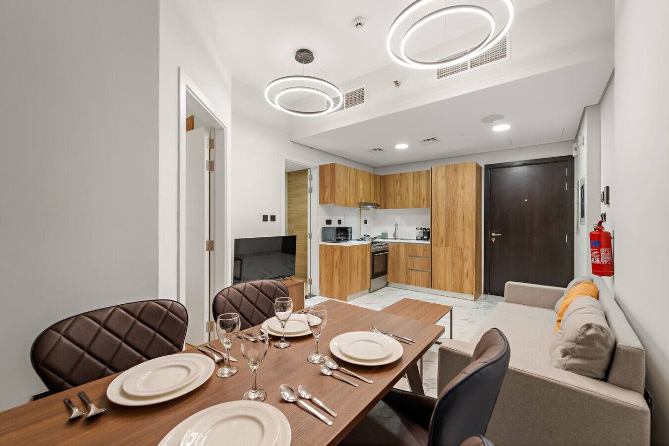 A modern dining area with a wooden table set for four, a kitchen in the background, and a fire extinguisher on the wall.
