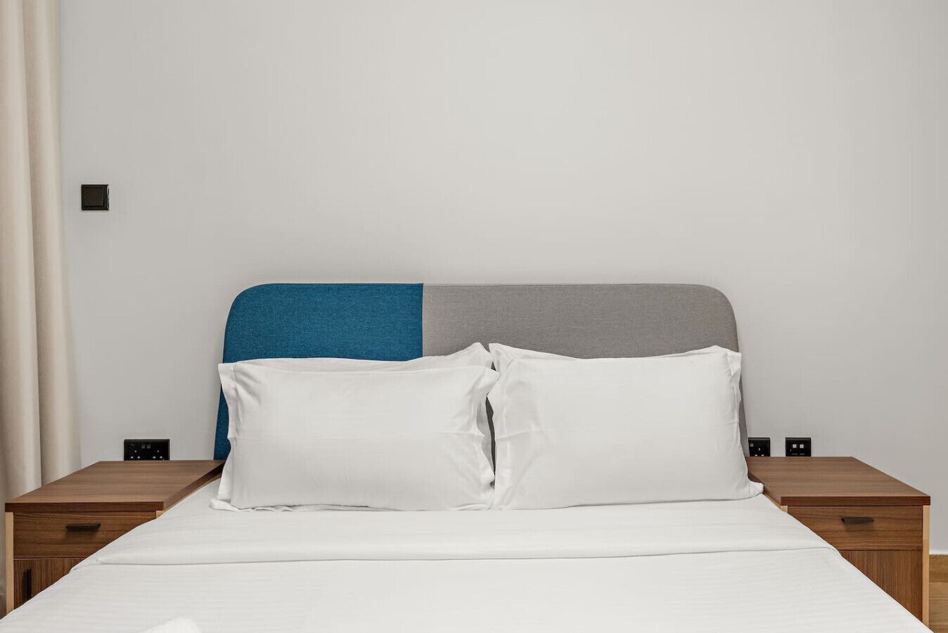 Ein Bett mit weißer Bettwäsche, einem blau-grauen Kopfteil und zwei Nachttischen mit schwarzen Schaltern.