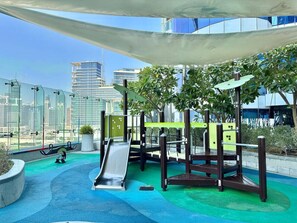 An outdoor playground with a slide and climbing structures, surrounded by greenery and a cityscape view.