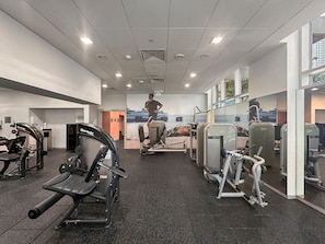 Uma academia com diversos equipamentos de exercício, uma pessoa caminhando e vista para o mar através de grandes janelas.