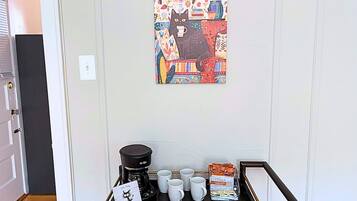A coffee station with a coffee maker, cups, and a coffee bag on a tray against a wall with a colorful painting.