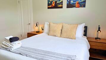 A neatly made bed with a white comforter and pillows, a bedside table with a lamp, and two paintings on the wall.