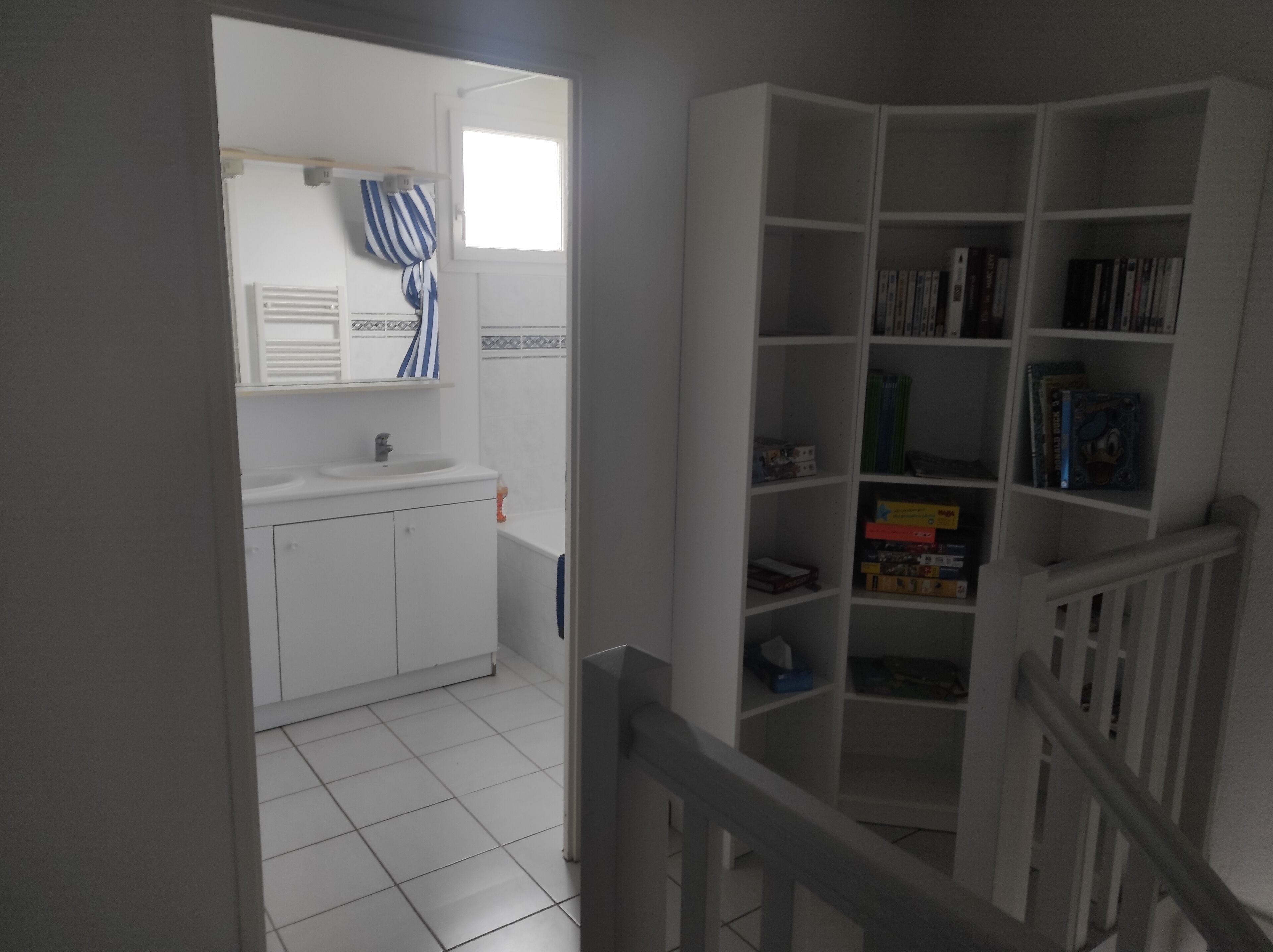 A bathroom with a white sink, mirror, and striped towel. Adjacent is a white bookshelf with various books and items.