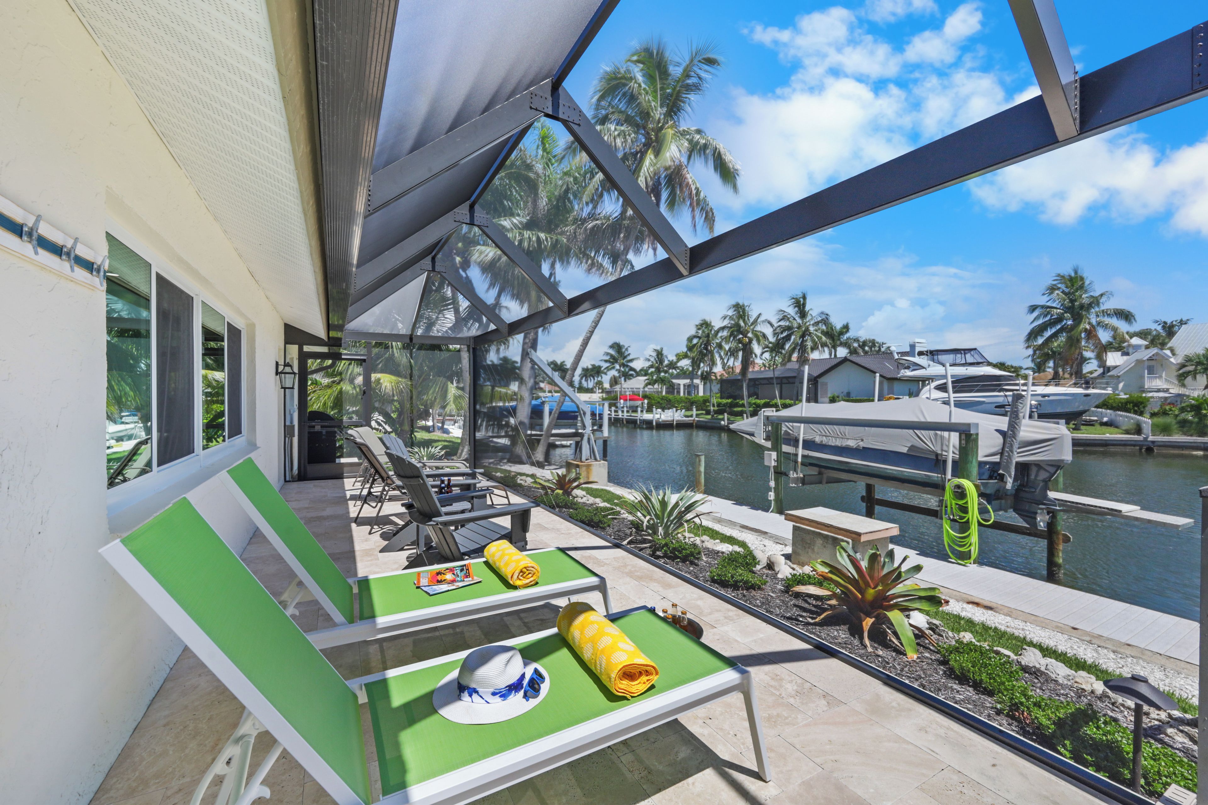 A covered outdoor seating area with green chairs and tables, overlooking a marina with boats and palm trees.