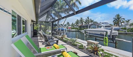 A covered outdoor seating area with green chairs and tables, overlooking a marina with boats and palm trees.