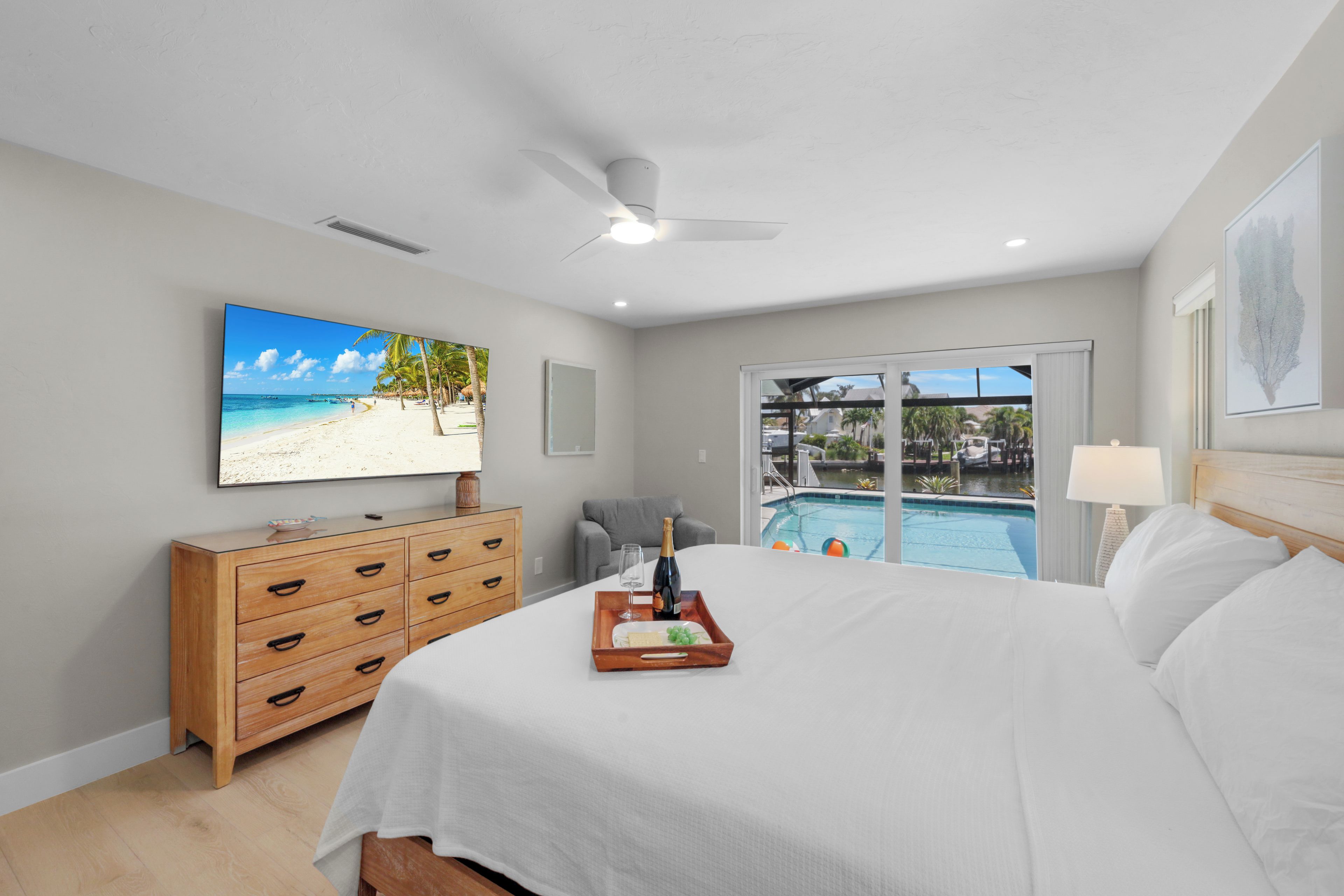 Une chambre avec un lit, une commode en bois, un ventilateur de plafond, une grande fenêtre donnant sur une piscine et des palmiers, et un tableau encadré accroché au mur.