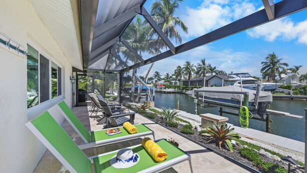 A covered outdoor seating area with green chairs and tables, overlooking a marina with boats and palm trees. - Canal home w/ pool, kayak, foosball, and ping pong (Marco Island)