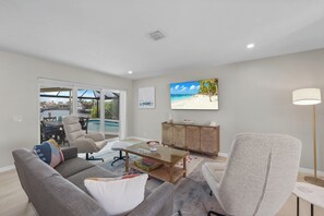 A modern living room with a glass coffee table, grey sofas, a wooden cabinet, and a large TV displaying a beach scene. - Canal home w/ pool, kayak, foosball, and ping pong (Marco Island)