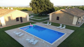 Ein rechteckiger Swimmingpool mit einem schwimmenden rosa Flamingo, umgeben von einer gepflasterten Liegefläche. Auf der Liegefläche stehen weiße Liegestühle und ein kleiner Tisch. Der Pool wird von einer niedrigen Hecke gesäumt, im Hintergrund ist ein großer Baum zu sehen.