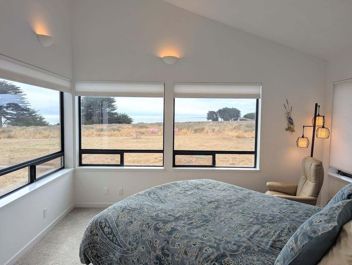 A bedroom with a large bed, two windows, a chair, and a wall-mounted lamp.