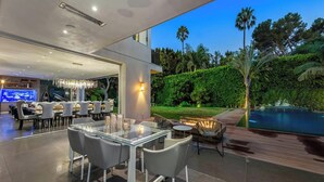 A modern outdoor dining area with glass tables and chairs, a pool, and a well-maintained garden with palm trees. - Alta Royale - Exquisite Designer Retreat Near Soho House & Rodeo Drive (West Hollywood)