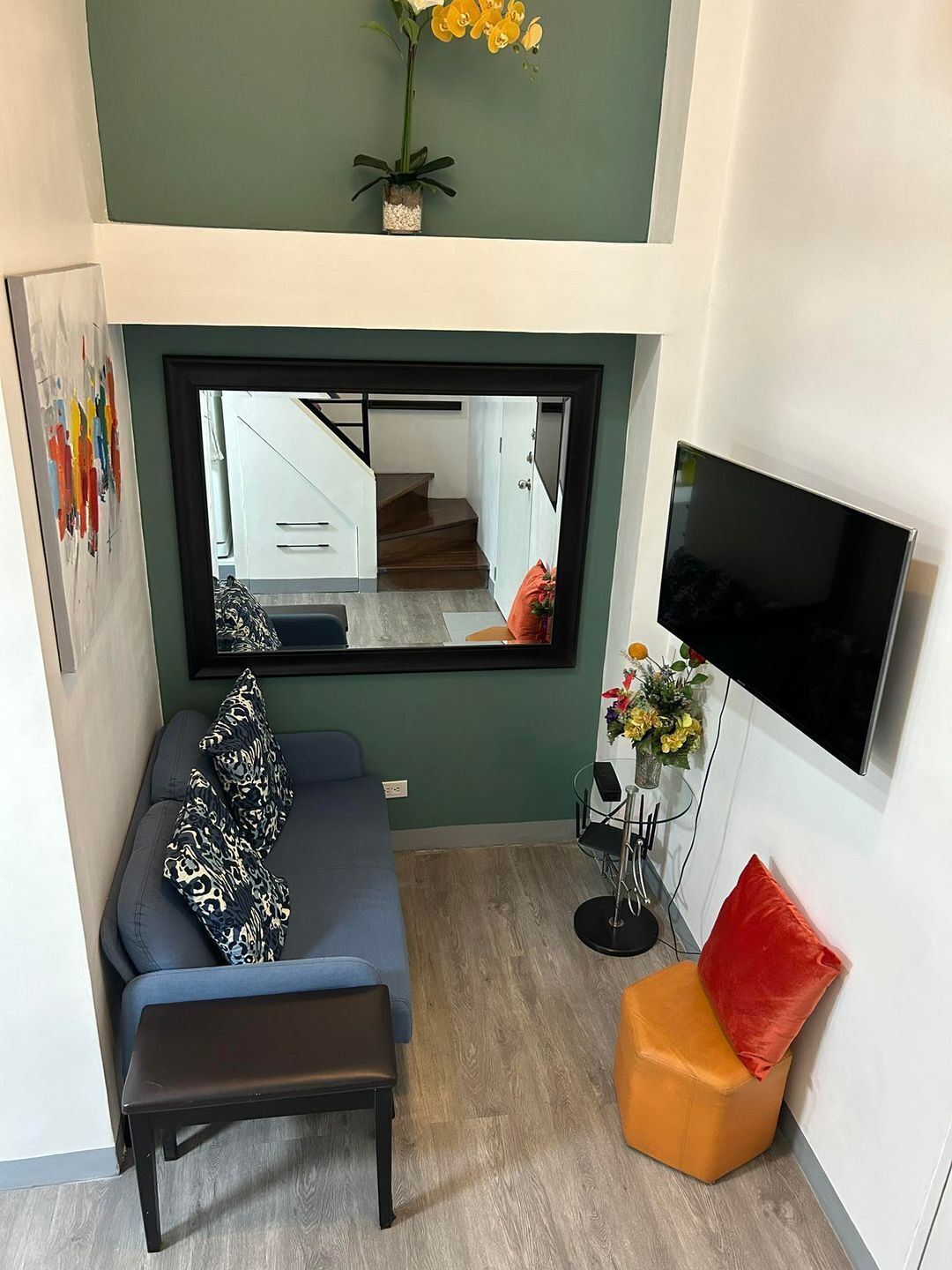 A modern living room with a sofa, a bench, a small orange ottoman, a mirror, a TV, and a flower arrangement.