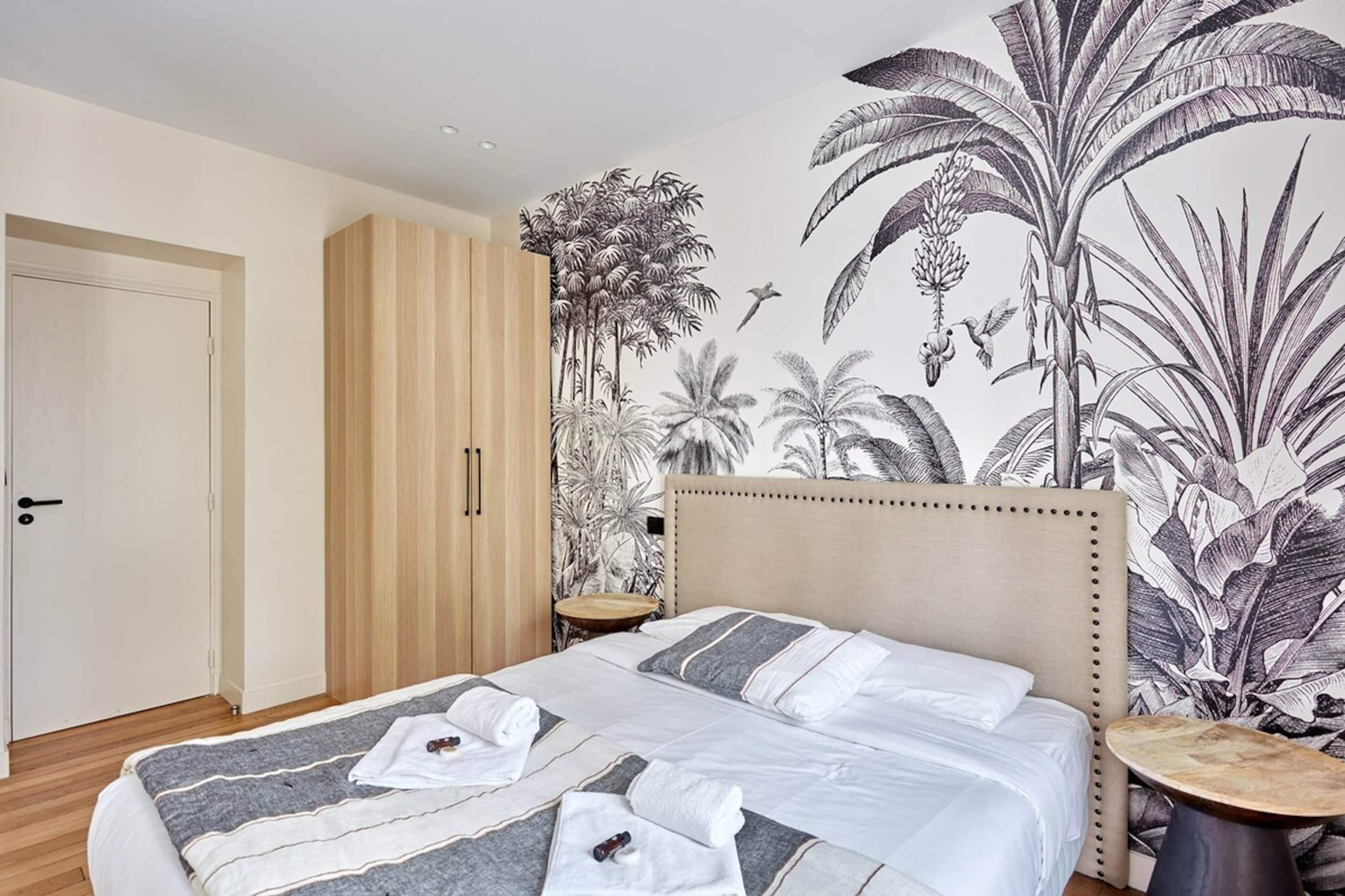 Un dormitorio con una cama grande, un armario de madera, una mesita redonda y un mural en la pared con plantas tropicales.