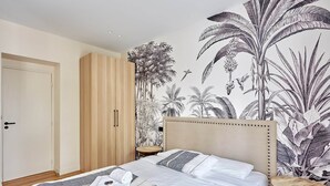 Ein Schlafzimmer mit einem großen Bett, einem hölzernen Kleiderschrank, einem kleinen runden Tisch und einer Wandmalerei mit tropischen Pflanzen.