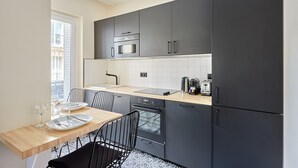 Eine moderne Küche mit dunklen Schränken, einer Mikrowelle, einem Ofen und einem Spülbecken. Ein Esstisch mit Stühlen und ein Fenster sorgen für natürliches Licht.