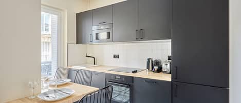 A modern kitchen with dark cabinets, a microwave, oven, and a sink. There is a dining table with chairs and a window providing natural light.
