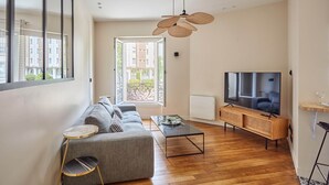 Ein modernes Wohnzimmer mit einem grauen Sofa, einem Couchtisch, einem Fernseher und einem großen Fenster mit Blick auf Gebäude.