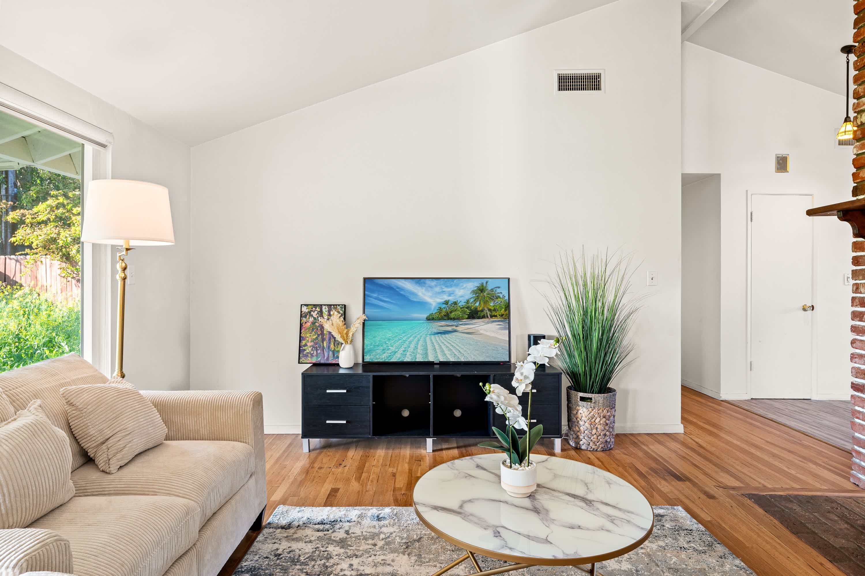 A modern living room with a beige sofa, a black entertainment unit, a large flat-screen TV, and a marble coffee table.