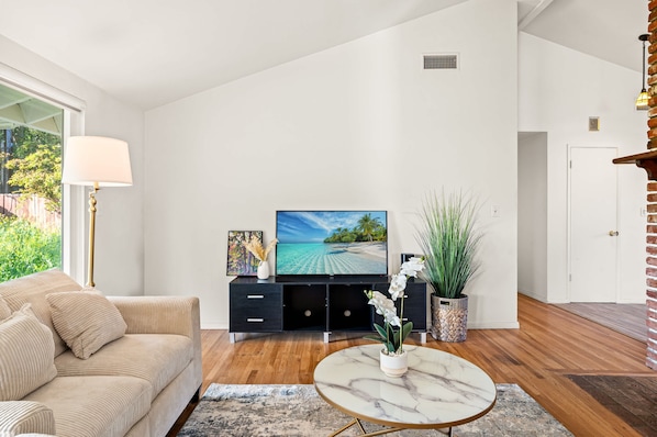 A modern living room with a beige sofa, a black entertainment unit, a large flat-screen TV, and a marble coffee table.