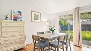 Un coin repas avec une table et des chaises en bois, une commode blanche avec des tiroirs, un tableau encadré au mur et une vue à travers une porte vitrée.