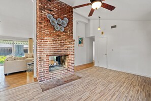 A spacious living room with a brick fireplace, wooden flooring, and a ceiling fan.