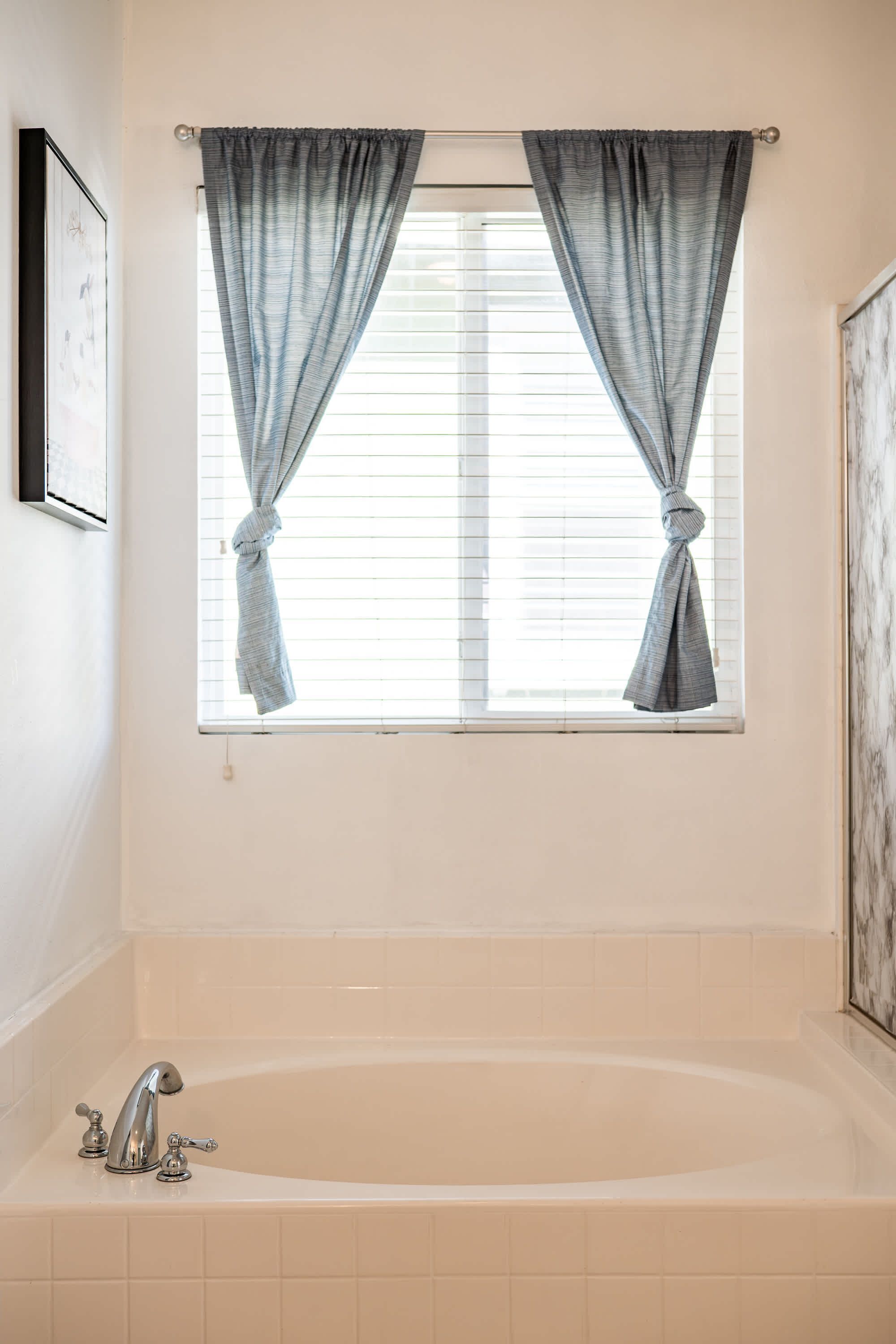 Un baño con bañera, una ventana con cortinas e imagen colgada en la pared.